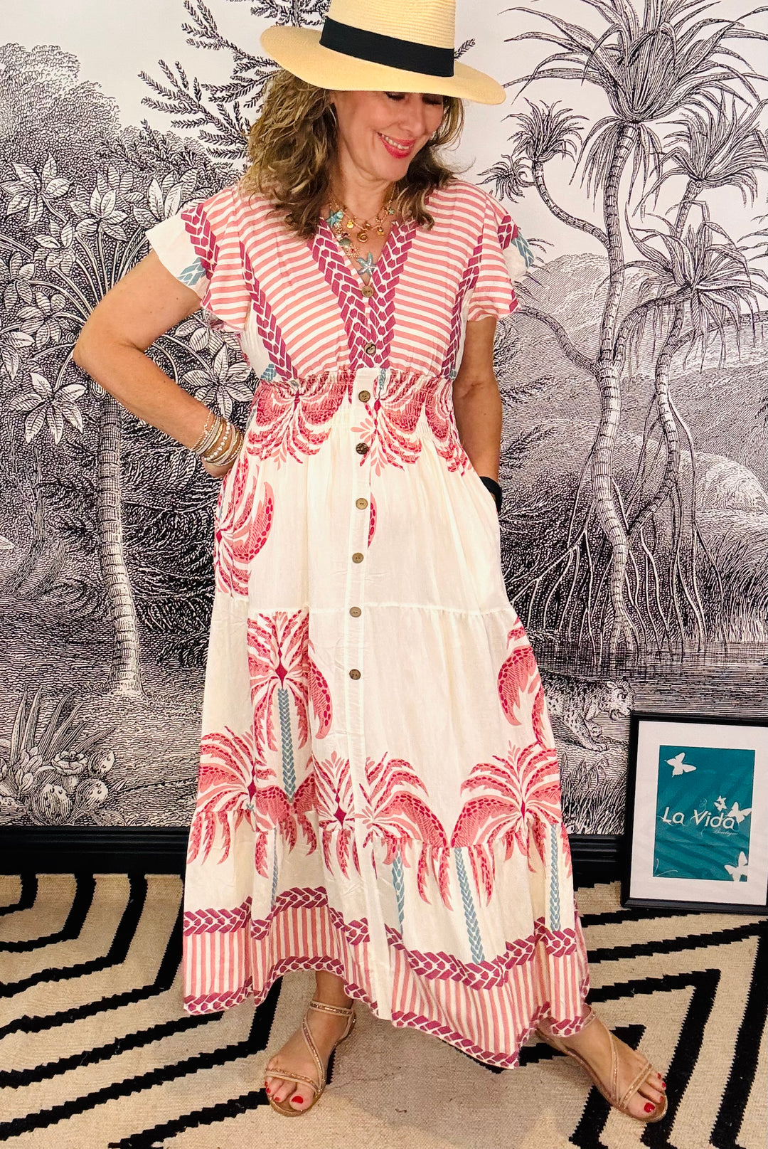 Donna Camera, founder of La Vida Boutique, wearing a red and white patterned dress with a straw hat in front of a decorative wall.
